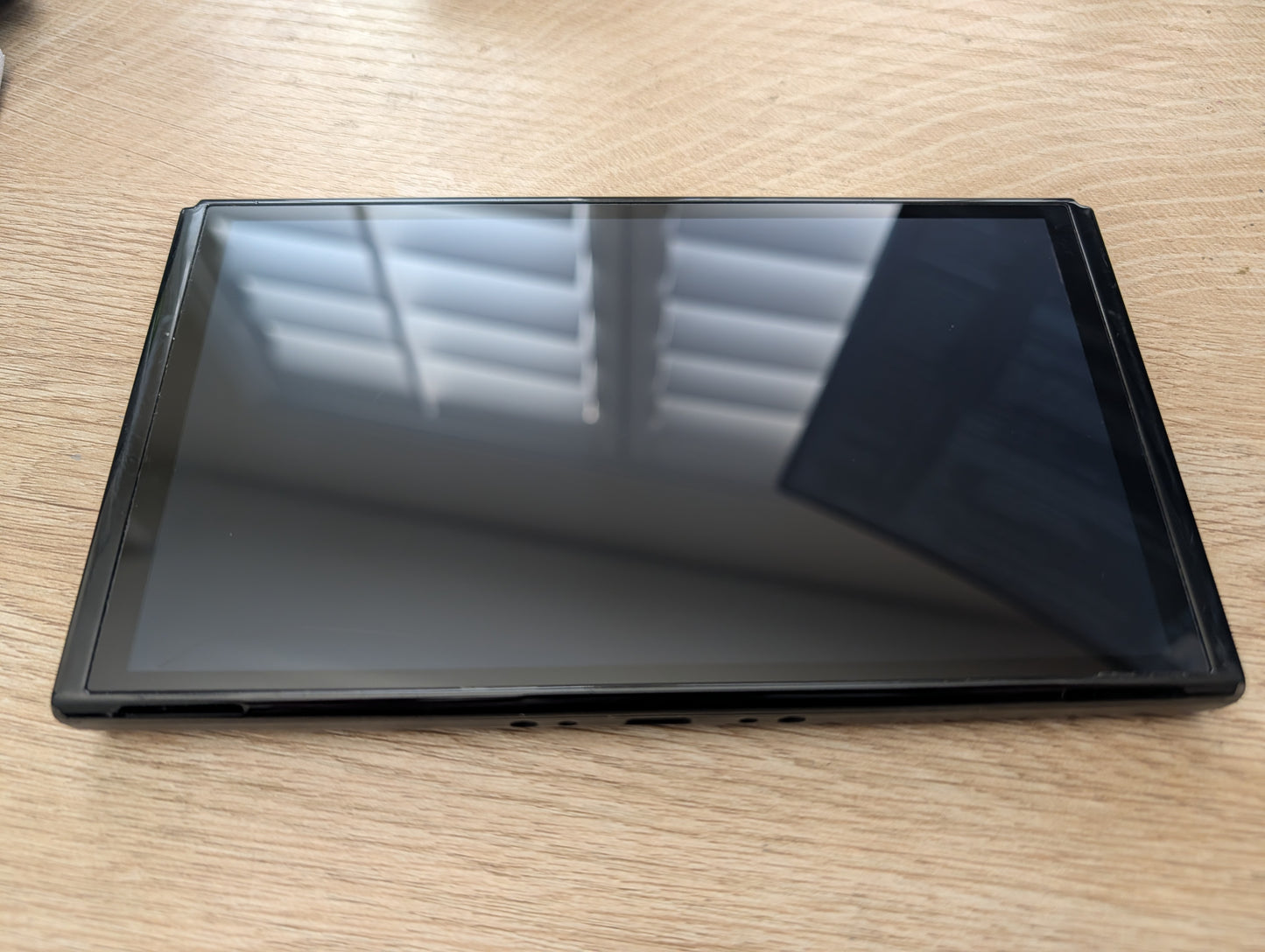 A black tablet or e-reader device with a glossy screen, lying flat on a light wood grain tabletop.