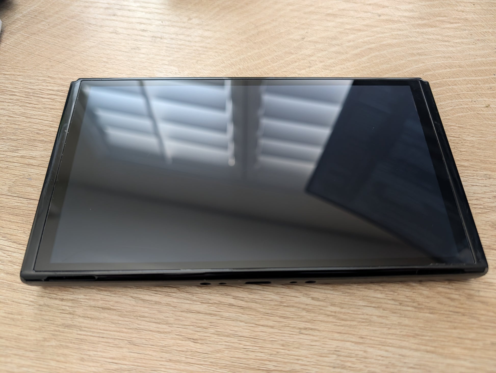 A black tablet or e-reader device with a glossy screen, lying flat on a light wood grain tabletop.