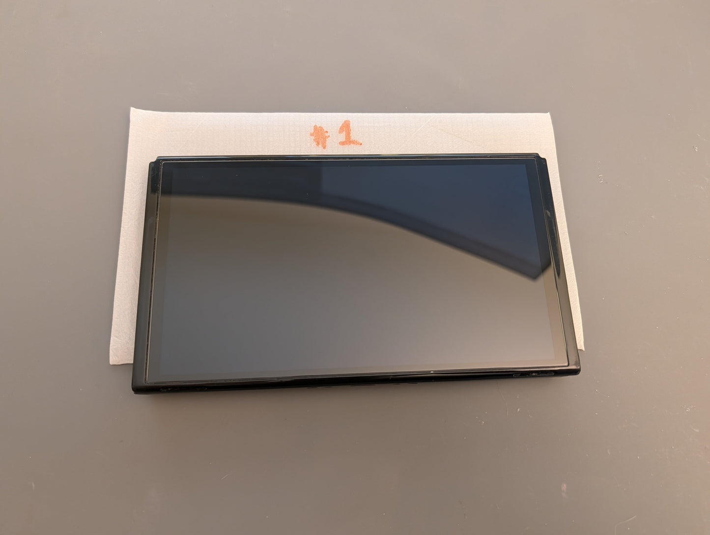 A dark gray tempered glass screen protector with a black frame, resting on a white protective paper backing with orange printed characters, set against a plain gray background.