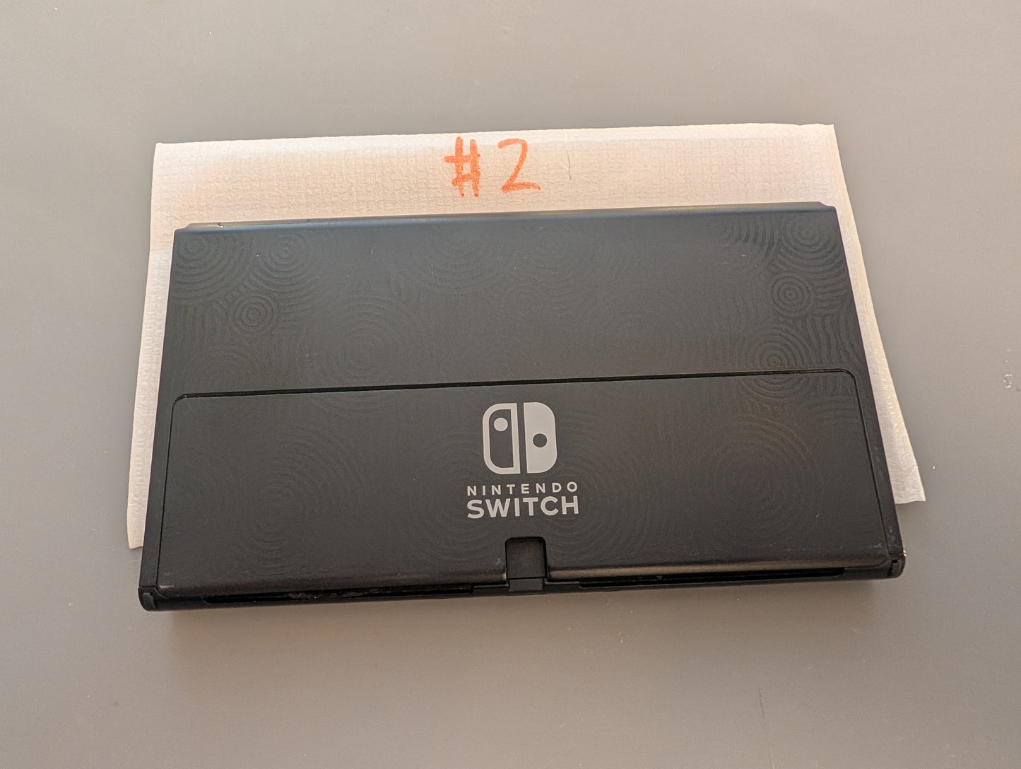 A black Nintendo Switch handheld console shown closed, placed on top of a white textured paper sheet with orange handwritten text on a gray surface.