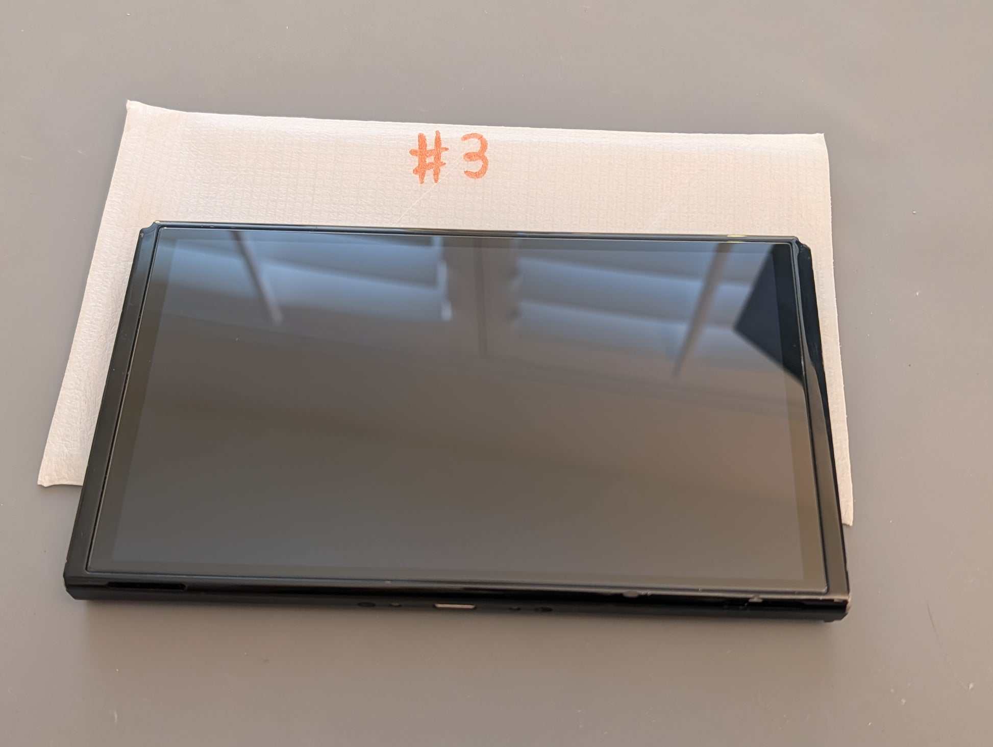 A black replacement screen panel for a tablet, labeled with a white protective sleeve marked '#3', on a gray surface.